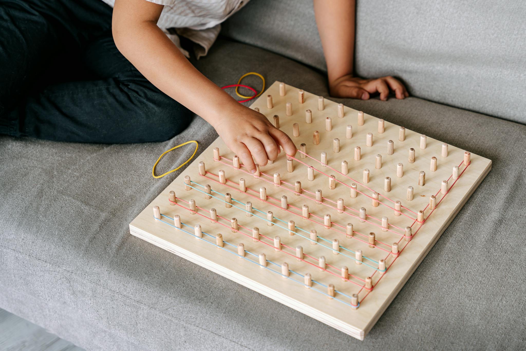 A child playing with a wooden pegboard, manipulating colorful rubber bands on a sofa for fun learning.